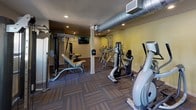 This single-floor fitness center is designed to provide a comprehensive workout environment with a practical and functional layout. The space is outfitted with a solid selection of cardio equipment, including multiple treadmills and elliptical machines from Matrix, positioned near windows that offer views of the surrounding outdoor area, allowing natural light to filter into the room. On the strength-training side, the facility includes cable machines, a lat pulldown station, a leg press, and a flat workout bench, offering a well-rounded range of resistance exercises. The flooring throughout consists of durable, patterned carpet tiles in earthy brown tones, lending a structured yet approachable feel to the space. Recessed lighting provides consistent illumination across the room, while an exposed ventilation duct runs prominently along the ceiling, contributing an industrial character to the otherwise neutral interior. The walls are finished in warm beige and soft gray tones. A wall-mounted flat-screen television is positioned for visibility across the exercise area. A dedicated exit door with exterior windows and emergency signage ensures safety and accessibility. The overall ambiance is clean, utilitarian, and well-maintained, making it a capable facility for a variety of fitness routines.