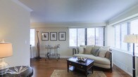 This single-floor space showcases a well-appointed living room designed with a classic, refined aesthetic. The room features warm hardwood flooring throughout, complemented by a textured ceiling and clean crown molding that frames the space with architectural detail. Large windows fitted with horizontal blinds line one wall, allowing abundant natural light to fill the room while offering elevated views of the surrounding cityscape — a notable feature that adds a dynamic urban backdrop to the interior.

The living area is anchored by a cream-toned sectional sofa dressed with an assortment of coordinating throw pillows, positioned around a dark-stained wood coffee table with drawer storage and decorative metallic accents. A patterned area rug grounds the seating arrangement, adding warmth and texture underfoot. A sleek metal bar cart positioned along an adjacent wall serves as a functional accent piece, while a trio of framed black-and-white prints arranged symmetrically above it adds a cohesive, gallery-like touch. A decorative tall giraffe figurine stands nearby, contributing a whimsical yet sophisticated element to the overall design. The space presents a balanced combination of comfort and style.