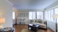 This single-floor space showcases a well-appointed living room designed with a classic, refined aesthetic. The room features warm hardwood flooring throughout, complemented by a textured ceiling and clean crown molding that frames the space with architectural detail. Large windows fitted with horizontal blinds line one wall, allowing abundant natural light to fill the room while offering elevated views of the surrounding cityscape — a notable feature that adds a dynamic urban backdrop to the interior.

The living area is anchored by a cream-toned sectional sofa dressed with an assortment of coordinating throw pillows, positioned around a dark-stained wood coffee table with drawer storage and decorative metallic accents. A patterned area rug grounds the seating arrangement, adding warmth and texture underfoot. A sleek metal bar cart positioned along an adjacent wall serves as a functional accent piece, while a trio of framed black-and-white prints arranged symmetrically above it adds a cohesive, gallery-like touch. A decorative tall giraffe figurine stands nearby, contributing a whimsical yet sophisticated element to the overall design. The space presents a balanced combination of comfort and style.