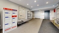 Located at 55 Bay St in Victoria, BC, this building lobby hallway serves as the primary entry and mail collection point for residents. The space is finished to a modern standard, combining large-format tile flooring near the entrance with carpet runner sections leading toward the elevator and inner corridors. Recessed ceiling lighting provides consistent, even illumination throughout.

A prominent feature is the expansive wall-mounted resident mailbox unit, organized in a grid format with individual labeled compartments. Alongside it sits a Canada Post parcel locker station, offering secure package pickup. A sleek upholstered bench provides a convenient waiting spot near the mailboxes, while a framed artwork piece adds a decorative touch to the neutral grey walls.

Toward the elevator area, a striking marble-effect accent wall panel introduces an element of elegance, with its white and grey veining creating a refined focal point. The corridor leading from the lobby connects to the elevator and building interior, lined with carpeting and finished with clean, minimal detailing. Safety features including a fire alarm panel and security camera are visibly integrated. The overall ambiance is clean, contemporary, and functional, reflecting a well-maintained residential building common area.