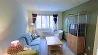 This single-floor space features a compact yet functional living area designed for comfortable daily use. The living room serves as the central gathering space, highlighted by a large window adorned with floral and dark curtain panels that frame views of surrounding trees, allowing ample natural light to brighten the room. The walls are painted in a combination of soft cream and muted sage green, creating a calm, welcoming atmosphere. Neutral-toned carpet runs throughout, adding warmth underfoot. The room comfortably accommodates a light blue sofa and a matching armchair, arranged around a simple wooden coffee table with a lower shelf for storage. A wall-mounted unit houses a television alongside a wooden media cabinet, offering practical storage and entertainment solutions. A floor lamp in the corner provides additional ambient lighting. A through-the-wall air conditioning and heating unit beneath the window ensures year-round climate control. An open metal shelving unit on the accent wall provides display and storage space. Overall, the space presents a modest, livable environment with a relaxed, homey character suited for everyday comfort.