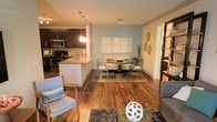 This single-story space showcases an open-concept layout that seamlessly connects the kitchen, dining, and living areas, creating a cohesive and functional flow. The kitchen features dark espresso cabinetry, stainless steel appliances, a subway tile backsplash, and a granite-topped island that doubles as a casual breakfast bar, illuminated by elegant pendant lighting overhead. The soft blue-grey accent wall in the dining area provides a calm, neutral backdrop for the round pedestal dining table surrounded by sleek acrylic and upholstered chairs, anchored by a patterned area rug. Decorative wall art and sculptural gold leaf accents add character to the dining space. Rich, warm-toned hardwood flooring extends throughout, tying each zone together harmoniously. The living area, partially visible in the foreground, features a glass coffee table and plush seating arranged on a textured area rug. Recessed and track lighting throughout ensures ample illumination, complementing the warm, inviting ambiance of the space. The overall aesthetic balances modern and transitional design elements for a polished, comfortable environment.