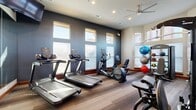 This dedicated exercise room is a compact yet thoroughly outfitted fitness facility designed to support a wide range of workout routines. The space features a striking design with dark charcoal accent walls contrasted against lighter upper walls, warm wood-tone flooring with subtle striped detailing, and recessed lighting complemented by a ceiling fan for air circulation.

Natural light floods the room through multiple windows positioned at both standard and elevated heights, creating an open, energizing atmosphere. The windows are fitted with horizontal blinds, allowing for adjustable light control throughout the day.

The room is equipped with an impressive array of fitness equipment, including multiple treadmills, an elliptical trainer, a stationary bike, and a rowing machine for comprehensive cardio options. A full multi-station cable resistance machine anchors the strength-training area, while a dumbbell rack stocked with free weights and a medicine ball rack add further versatility. Wall-mounted flat-screen televisions provide entertainment during workouts.

Full-length mirrors on one wall visually expand the space and assist with form during exercise. A fire safety station and posted rules signage reflect attention to safety standards. The overall ambiance is modern, functional, and motivating — well-suited for a dedicated fitness environment.