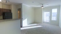 This single-floor residence offers a clean, open layout designed for comfortable everyday living. The main living area is a standout feature, bathed in abundant natural light that pours in through large windows dressed with blinds, casting warm patterns across the plush wall-to-wall carpeting. Crown molding frames the room with a refined architectural detail, while a ceiling fan with a light fixture provides both comfort and practical illumination overhead. The neutral, light-toned walls create a versatile backdrop that enhances the sense of openness throughout the space. A partial countertop or breakfast bar transitions seamlessly from the living area toward the kitchen zone, hinting at an open-concept design that encourages easy flow between spaces. A doorway visible from the main living area leads to an additional room, suggesting a well-connected and functional floor plan. The overall ambiance is bright, airy, and inviting, with a move-in-ready feel characterized by its clean finishes and thoughtfully designed layout. The combination of natural light, soft carpeting, and classic architectural details gives this residence a warm and welcoming character.