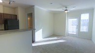 This single-floor residence offers a clean, open layout designed for comfortable everyday living. The main living area is a standout feature, bathed in abundant natural light that pours in through large windows dressed with blinds, casting warm patterns across the plush wall-to-wall carpeting. Crown molding frames the room with a refined architectural detail, while a ceiling fan with a light fixture provides both comfort and practical illumination overhead. The neutral, light-toned walls create a versatile backdrop that enhances the sense of openness throughout the space. A partial countertop or breakfast bar transitions seamlessly from the living area toward the kitchen zone, hinting at an open-concept design that encourages easy flow between spaces. A doorway visible from the main living area leads to an additional room, suggesting a well-connected and functional floor plan. The overall ambiance is bright, airy, and inviting, with a move-in-ready feel characterized by its clean finishes and thoughtfully designed layout. The combination of natural light, soft carpeting, and classic architectural details gives this residence a warm and welcoming character.