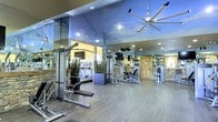 This fitness center is a thoughtfully designed space that combines functionality with an inviting aesthetic. The vaulted ceiling, adorned with a sky-inspired mural, creates an open and airy atmosphere, while a large industrial ceiling fan ensures comfortable airflow throughout the facility. Floor-to-ceiling mirrors line the walls, amplifying the sense of space and allowing members to monitor their form during workouts. The flooring consists of durable, hardwood-style planks that lend a clean and modern look to the space. Rustic stone accent walls add a warm, natural contrast to the contemporary equipment and mirrored surfaces. The fitness center is outfitted with a comprehensive selection of equipment, including cable and resistance strength machines, adjustable benches, and cardio machines such as treadmills. Recessed lighting and mounted TVs are positioned throughout the space for convenience and entertainment. Decorative planters with greenery are placed strategically, adding a touch of nature to the environment. The overall ambiance strikes a balance between a high-end, resort-style feel and a practical, fully functional fitness facility suited for a range of workout routines.