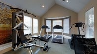 This dedicated fitness room offers a thoughtfully designed workout space combining both functionality and architectural appeal. The room's most striking feature is its vaulted ceiling, which soars to a sharp peak and is fitted with recessed lighting, creating an open, airy atmosphere that enhances the sense of space during exercise.

The walls are finished in a warm beige tone, complemented by white trim and baseboards that provide clean contrast throughout. A distinctive curved bay window alcove anchors one end of the room, fitted with white blinds that allow for adjustable natural light. Additional windows along the side wall further brighten the space while maintaining privacy.

The fitness area is outfitted with two side-by-side treadmills positioned near the bay window, offering a pleasant focal point during cardio workouts. A multi-function strength training cable machine with an adjustable bench occupies the foreground, providing a range of resistance exercise options. Additional equipment is visible along the perimeter, rounding out the room's fitness capabilities.

Neutral carpet covers the floor throughout, with rubber matting placed beneath the cardio equipment for protection and stability. The overall layout is efficient and well-organized, making it a practical and inviting space for daily exercise.