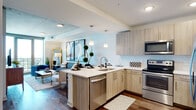 This single-floor unit showcases a thoughtfully designed open-concept layout that seamlessly connects the kitchen, dining, and living areas. The kitchen is outfitted with light wood cabinetry, white quartz countertops, a mosaic tile backsplash, and a full suite of stainless steel appliances including a range, over-the-range microwave, dishwasher, and refrigerator. A peninsula with bar seating and pendant lighting above serves as the centerpiece, offering both additional prep space and casual dining. Rich hardwood flooring flows throughout the main living areas, grounding the contemporary aesthetic.

The living room is bathed in natural light from floor-to-ceiling windows and provides direct access to a private balcony, offering sweeping panoramic views of the surrounding cityscape and landscape. An exposed concrete ceiling adds an industrial contrast to the otherwise clean, modern finishes.

The bedroom continues the theme of abundant natural light with large corner windows framing expansive outdoor views and balcony access. The bathroom is finished with large-format marble-style tile flooring, a wood-toned vanity with a quartz countertop, a framed mirror, and a glass-enclosed walk-in shower, creating a clean, spa-like atmosphere throughout.