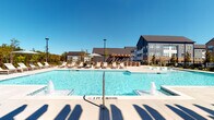 The Atwater pool amenity area in Oakwood, Georgia is a thoughtfully designed outdoor retreat that combines modern aesthetics with relaxed, resort-inspired living. At the heart of the space is a generously sized swimming pool with crystal-clear blue water, accented by built-in fountain jets that add a dynamic visual element. The pool deck is finished in smooth, light-colored concrete, providing ample space for lounging and socializing under the open Georgia sky.

A row of chaise lounge chairs lines the poolside, offering residents dedicated spots to relax and soak in the sun. A large market umbrella provides shaded seating for those seeking relief from the sun. The standout architectural feature of the space is a substantial wood-beam pergola with rustic timber posts, draped with string lights that lend a warm, inviting ambiance during evening hours. Beneath the pergola, a comfortable seating arrangement creates a shaded gathering area adjacent to the pool.

The pool house building, finished in crisp white board-and-batten siding with dark-framed glass doors and windows, serves as a functional anchor to the space. Surrounding the area, views of the multi-story residential buildings and mature treeline provide a picturesque backdrop. The space is enclosed by a black iron fence, ensuring a secure and well-defined environment for residents to enjoy year-round.