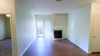 This single-floor space centers around a well-appointed living area that combines practical design with comfortable features. The room is anchored by a tile-surround fireplace with a classic white mantel, serving as a natural focal point along the main wall. The fireplace's clean lines and neutral tiling complement the overall aesthetic of the space.

Rich hardwood flooring extends throughout the living area, adding warmth and a sense of continuity to the room. The floors are in good condition, offering a durable and visually appealing foundation for the space.

A sliding glass door, dressed with vertical blinds, provides direct access to an outdoor area and allows natural light to filter into the room. The glimpse of an exterior fence and greenery beyond suggests a modest private outdoor space.

The walls are finished in a neutral off-white tone, keeping the space bright and versatile. A textured popcorn ceiling adds a layer of acoustic character typical of the architectural era. Visible ceiling vents indicate a central HVAC system for year-round climate control. White baseboards frame the perimeter, providing a clean finish throughout the living area.