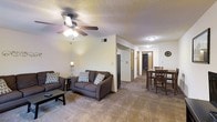 This single-floor residential space offers a welcoming and functional layout designed for comfortable everyday living. The open-concept main area seamlessly connects the living room and dining space, creating a cohesive and practical flow throughout.

The living room is anchored by a plush, dark-toned sofa and loveseat set, accented with patterned throw pillows that add a touch of personality to the neutral color palette. A ceiling fan with an integrated light fixture provides both overhead illumination and air circulation. Soft carpeting underfoot enhances the warmth of the room, while a modest coffee table rounds out the seating area.

Adjacent to the living room, the dining area features a classic wooden table and chair set, ideal for everyday meals. The space transitions smoothly into a hallway leading to additional rooms, with wood-trimmed doorways adding a subtle traditional accent throughout.

The textured ceiling and neutral beige walls contribute to a calm, understated ambiance. Recessed lighting in the hallway ensures the space remains well-lit beyond the main living area. Overall, this unit offers a clean, move-in-ready environment with a straightforward, functional design suited for comfortable residential living.
