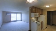 This single-floor space presents an open-concept design that integrates the kitchen and living area into one cohesive layout. The kitchen is equipped with white appliances, including an electric coil range, an over-the-range microwave, and a full-size refrigerator. Medium-toned wood cabinetry offers ample overhead storage, while the countertop provides functional workspace. Under-cabinet lighting adds a practical touch to the cooking area.

The living area extends from the kitchen, featuring wall-to-wall carpet in a neutral blue-gray tone that lends a soft, comfortable feel to the space. A ceiling fan with a light fixture serves as the central overhead lighting source, contributing to both air circulation and ambient illumination.

A large sliding glass door fitted with vertical blinds dominates one wall of the living area, allowing generous natural light to enter and offering potential outdoor access. The textured ceiling finish is consistent throughout, and the neutral wall tones keep the space feeling open and adaptable. Overall, the layout is practical and straightforward, suited for everyday living.