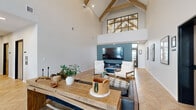 Located at 5532 38th St S in Fargo, North Dakota, this thoughtfully designed clubhouse delivers an impressive blend of modern farmhouse aesthetics and functional community space. The single-floor layout makes efficient use of its footprint, with an open-concept design that feels remarkably spacious thanks to dramatic vaulted ceilings adorned with exposed natural wood beam trusses. A striking black-framed clerestory window at the ceiling peak floods the interior with natural light while adding an architectural focal point.

The main lounge area is anchored by a sleek electric fireplace set against a deep teal accent wall, paired with a wall-mounted television and comfortable seating arrangements. A substantial reclaimed wood console table grounds the space and contributes to its warm, inviting character. Herringbone-patterned hardwood flooring runs throughout, adding texture and elegance underfoot.

Near the entrance, large black-framed glass doors and expansive windows bring in abundant natural light and offer views of the surrounding community. A reception or leasing desk area provides a functional workspace, while a dedicated refreshment station features white cabinetry with gold hardware, dark countertops, a subway tile backsplash, and a coffee service — ideal for welcoming residents and guests. The overall ambiance is polished yet comfortable, balancing sophistication with approachability.