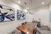 This well-appointed single-room space functions as both a dining and meeting room, offering a refined yet functional environment. The room is anchored by a striking live-edge wood dining table surrounded by plush gray upholstered chairs, providing comfortable seating for a small group. Overhead, a contemporary multi-globe pendant chandelier serves as a focal point, casting warm, ambient light throughout the space.

The flooring features a sophisticated herringbone pattern in neutral gray tones, adding texture and visual depth underfoot. Crisp white walls create a clean backdrop, enhanced by a pair of large-format abstract paintings rendered in bold shades of blue and white, lending the room an artistic, gallery-like quality.

A mounted teal glass whiteboard on one wall adds a functional, collaborative element, making the space well-suited for meetings or working sessions. A modern white interior door with glass panel insets provides access while maintaining an open, airy feel. Recessed ceiling lighting supplements the pendant fixture, ensuring the room is evenly and thoroughly illuminated. Thoughtful decorative accents, including a geometric terrarium and small floral arrangements, complete the polished aesthetic of this compact yet impressively detailed space.