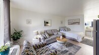 Located at 2003 Pearl St in Beaufort, South Carolina, this cozy coastal studio offers a thoughtfully arranged single-level layout that maximizes every inch of its compact footprint. The open-plan living area seamlessly integrates the sleeping space, sitting area, and kitchenette into one cohesive room, creating a functional and welcoming environment.

The interior is finished with light wood-style flooring throughout, complemented by crisp white walls adorned with beach-inspired artwork, including coastal landscape prints and nautical accents. Crown molding adds a refined touch to the living area, while a large window with dark curtain panels allows natural light to fill the space. The seating area features a plush gray sofa, an accent chair with a geometric blue-and-white pattern, and a rustic round wooden coffee table, all arranged around a flat-screen television on a white wicker media console.

The sleeping area is integrated naturally into the layout, offering a queen-sized bed with layered neutral bedding and decorative pillows. A wall-mounted mirror near the kitchenette nook enhances the sense of openness. The kitchenette includes white cabinetry, a stainless steel full-sized refrigerator, and a dedicated countertop workspace. An adjacent bathroom with vanity lighting and mirror completes the unit. The overall ambiance is relaxed and beach-cottage in character, ideal for Lowcountry living.
