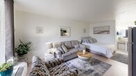 Located at 2003 Pearl St in Beaufort, South Carolina, this cozy coastal studio offers a thoughtfully arranged single-level layout that maximizes every inch of its compact footprint. The open-plan living area seamlessly integrates the sleeping space, sitting area, and kitchenette into one cohesive room, creating a functional and welcoming environment.

The interior is finished with light wood-style flooring throughout, complemented by crisp white walls adorned with beach-inspired artwork, including coastal landscape prints and nautical accents. Crown molding adds a refined touch to the living area, while a large window with dark curtain panels allows natural light to fill the space. The seating area features a plush gray sofa, an accent chair with a geometric blue-and-white pattern, and a rustic round wooden coffee table, all arranged around a flat-screen television on a white wicker media console.

The sleeping area is integrated naturally into the layout, offering a queen-sized bed with layered neutral bedding and decorative pillows. A wall-mounted mirror near the kitchenette nook enhances the sense of openness. The kitchenette includes white cabinetry, a stainless steel full-sized refrigerator, and a dedicated countertop workspace. An adjacent bathroom with vanity lighting and mirror completes the unit. The overall ambiance is relaxed and beach-cottage in character, ideal for Lowcountry living.