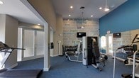 This fitness center is a thoughtfully designed workout facility that combines functionality with motivational aesthetics. The space features a neutral color palette accented by a bold teal-blue feature wall, creating an energizing atmosphere for exercise. Recessed lighting and ceiling fans ensure the space remains well-lit and comfortable during workouts.

A standout design element is the large motivational word-art mural covering the main wall, displaying phrases such as "strength," "determination," "inspire," and "lift," setting an encouraging tone throughout the space. The room is carpeted in a durable blue-gray tone suited for a fitness environment.

The facility is outfitted with a selection of strength-training machines, including a multi-function cable and resistance unit, as well as a dedicated weight bench with a rack of free weights and dumbbells. A wall-mounted flat-screen television provides entertainment during workouts. Natural light filters in through windows, contributing to an open and welcoming feel. A scale is also available near the entrance area.

Entry to the fitness center is through frosted glass double doors, offering both privacy and a clean, modern aesthetic. The overall design balances practicality and motivation, making it a well-rounded space for residents or members seeking a convenient and fully equipped place to exercise.