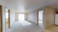 This single-floor residential space offers a clean, neutral aesthetic designed for comfortable everyday living. The main living area is generously proportioned, featuring soft wall-to-wall carpeting that adds warmth underfoot and contributes to an inviting atmosphere. Smooth white ceilings keep the space feeling open and airy, while the neutral gray and beige wall tones provide a versatile backdrop suitable for a variety of décor styles. A large window dressed with vertical blinds allows an abundance of natural light to pour into the living area, casting bright, even illumination across the room and creating a welcoming ambiance. A baseboard heating unit runs along the exterior wall, ensuring year-round comfort. Warm-toned wood door trim frames the passageway leading into an adjoining room, which also features carpeting and appears to benefit from its own natural light source. The overall layout flows naturally between spaces, offering a functional and cohesive living environment. The unit's simple, well-maintained finishes make it a practical and move-in-ready space.