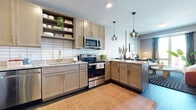This well-designed one-bedroom, one-bathroom unit is laid out across a single floor, making efficient use of its compact footprint. The open-concept kitchen and living area form the heart of the space, with the kitchen showcasing light taupe shaker-style cabinetry, granite countertops, a white subway tile backsplash, and a full suite of stainless steel appliances including a range, microwave, and refrigerator. Open shelving above the sink adds a modern, airy touch, while pendant lighting over the peninsula ties the kitchen seamlessly into the living area. The living room benefits from large windows that invite abundant natural light and offer views of the surrounding building exterior. The bedroom is a cozy retreat featuring carpeted flooring, a tray ceiling detail, and a sliding glass door opening to a private balcony. The bathroom is clean and functional, featuring a tub-shower combination, wood-look tile flooring, and gold-toned hardware accents. Dark hardwood-style flooring runs through the main living areas, grounding the warm, contemporary aesthetic carried throughout the unit.