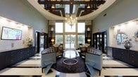 This striking single-floor space presents an impressive lobby defined by a dramatic circular rotunda ceiling adorned with dark wood geometric trim and an inset mirrored panel that visually expands the volume overhead. A sculptural, ornate chandelier serves as the centerpiece, suspended beneath the rotunda and drawing the eye upward to emphasize the soaring ceiling height. Floor-to-ceiling windows flanking the main entrance allow abundant natural light to pour in, offering clear sightlines to the outdoors and creating an airy, open atmosphere. The flooring features a bold black-and-white geometric tile pattern that anchors the space with a sense of classic elegance. A circular area rug defines the central seating arrangement, where curved, high-backed upholstered sofas and contemporary accent chairs invite comfortable conversation. A sleek glass-and-metal circular coffee table completes the seating vignette. Flanking lounge areas with warm lamp lighting and draped curtains create more intimate zones within the expansive layout. Dark-toned walls and cabinetry contrast beautifully with the lighter architectural elements, contributing to a refined, hotel-like ambiance throughout.