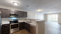 Located at 1025 Kensington Dr in Jacksonville, North Carolina, this single-floor residence offers a well-designed, open-concept layout that seamlessly connects the kitchen and living areas. The kitchen is a standout feature, equipped with stainless steel appliances — including a refrigerator, electric range, built-in microwave, and dishwasher — paired with rich, dark cabinetry, light countertops, a single-basin sink, and wood-look flooring. A peninsula counter creates a natural boundary between the kitchen and the expansive living area, which features the same durable wood-look flooring and textured ceilings throughout. The living room is generously sized and benefits from natural light through a large front-facing window with views of the surrounding neighborhood. Bedrooms are carpeted for comfort and feature windows that invite natural light. The bathrooms are thoughtfully finished, with one featuring a bold blue vanity with a white countertop and wood-look tile flooring, and another offering a full tub-shower combination, a dark wood vanity, and an adjacent walk-in closet with built-in shelving. Neutral wall tones and clean white trim create a cohesive, move-in-ready aesthetic throughout the entire space.
