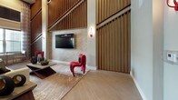 This single-floor hospitality space showcases a refined contemporary design that blends warmth and boldness throughout its open layout. The lounge area immediately draws attention with its dramatically tall ceilings clad in floor-to-ceiling vertical wood slat paneling, creating a rich textural backdrop that anchors the space. A wall-mounted flat-screen television is set against a subtly patterned accent wall, while a sculptural red chair and a low-profile coffee table atop a decorative area rug add a striking pop of color and artistic flair. Natural light filters in through tall windows fitted with blinds, brightening the light-toned wide-plank tile flooring that extends seamlessly throughout. A small bistro table and chairs near the windows offer an intimate seating nook.

The corridor connecting the spaces features arched openings accented by distinctive red pendant sconces, lending a sense of rhythm and visual continuity between zones. The bar and lounge service area is a standout feature, equipped with a long white quartz-topped island with barstool seating, open wood shelving, stainless steel appliances, a beverage refrigerator, and stylish black cone pendant lighting overhead. Fresh floral arrangements add vibrant touches of color. Together, the spaces project a polished, modern hospitality ambiance with thoughtful design details throughout.