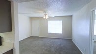 This single-floor space presents a straightforward and functional layout. The main living area is finished with wall-to-wall carpet in a neutral tone, contributing to a warm and quiet atmosphere underfoot. A ceiling fan with an integrated light fixture is centrally mounted, providing both air circulation and ambient lighting throughout the room. The textured popcorn ceiling adds a classic finishing detail overhead. A single window fitted with horizontal blinds allows natural light to filter into the space, brightening the neutral-painted walls. A white interior door is positioned along one wall, providing access to an adjacent area or entry point. White baseboards frame the perimeter of the room, offering a clean contrast against the carpeting and walls. The overall color palette is understated, consisting of soft beige and white tones that create a calm, neutral environment. The space is open and unobstructed, offering flexibility in furniture arrangement and use.