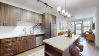 This thoughtfully designed single-floor residence offers a seamless blend of contemporary style and functional living. The open-concept kitchen serves as a central highlight, featuring rich dark wood cabinetry, white quartz countertops, a subway tile backsplash, and a full suite of stainless steel appliances including a refrigerator, dishwasher, range, and over-the-range microwave. A central kitchen island with built-in shelving provides additional prep space and storage, while pendant and track lighting add both ambiance and practicality. Wood-look flooring flows throughout the main living areas, complementing the warm, modern aesthetic. The living room is bathed in natural light from multiple large windows with horizontal blinds, creating a bright and welcoming atmosphere. A glass door offers access to the outdoors, further enhancing the sense of openness. The bedroom features plush carpeting, a ceiling fan, a bold accent wall in a soft blue tone, and generous natural light from dual windows. Additional bedrooms offer comparable comfort with ample closet access. The bathroom is finished with a dark wood vanity, quartz countertop, framed mirror, and lighted vanity fixtures, delivering a clean, upscale feel throughout.