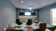 This single-floor space is designed as a contemporary lounge or media room, thoughtfully appointed for both comfort and functionality. The room's most prominent feature is a wall of three flat-screen televisions — a large central display flanked by two smaller screens — mounted above a continuous run of built-in cabinetry with clean-lined drawers and a light countertop surface. Wall-mounted speakers on either side of the display wall provide an integrated audio experience, making this an ideal setting for presentations, screenings, or casual viewing.

The interior is finished in a muted blue-gray palette that lends the space a calm, professional atmosphere. Recessed ceiling lighting is complemented by a central ceiling fan, ensuring the room stays well-lit and comfortable. Rich hardwood-style flooring in a herringbone pattern adds warmth and visual interest underfoot, providing a refined contrast to the cool wall tones.

Seating is arranged in a conversational cluster at the center of the room, featuring modern curved lounge chairs with sleek metallic bases and plush upholstery in deep teal and soft gray tones. A small round accent table anchors the seating arrangement. The overall ambiance is polished and inviting, balancing media functionality with a relaxed, social atmosphere.
