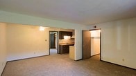 This single-floor residential unit offers a practical and functional layout designed for comfortable everyday living. The main living area is generously sized, featuring neutral wall-to-wall carpeting that adds warmth underfoot, white walls that contribute to a bright and open feel, and a ceiling fan that provides both air circulation and ambient lighting. The space flows naturally into the kitchen, which is positioned centrally and outfitted with dark wood cabinetry, a countertop sink, and under-cabinet lighting that creates a warm, inviting atmosphere. White appliances, including a refrigerator and dishwasher, complement the practical nature of the kitchen. A doorway from the living area leads to a closet or storage space, offering useful organization options. A hallway visible from the main living space connects to additional rooms, suggesting a well-thought-out floor plan that separates living and private areas effectively. The overall interior features consistent neutral tones, standard-height ceilings, and straightforward finishes throughout, making it a clean and move-in-ready space suited for a range of occupants.