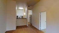 This single-floor space is designed with an open, flowing layout that promotes a sense of connectivity between its main living areas. The kitchen is positioned along one side and features a dark-topped breakfast bar counter, white cabinetry, stainless steel appliances including a refrigerator and microwave, and recessed overhead lighting. The counter-height bar creates a natural division between the kitchen and the adjoining living area without fully closing off the space.

The flooring throughout is warm-toned hardwood-style, providing a cohesive and clean foundation across the main areas. Walls are finished in a soft neutral tone, contributing to a bright and airy atmosphere. Notably elevated ceilings add a sense of verticality and openness to the space, enhancing the overall roominess of the interior.

An arched passageway is visible toward the rear of the main hallway, leading deeper into the unit. Additional rooms are accessible via white panel doors, offering privacy while maintaining the clean aesthetic of the space. The layout appears well-organized, with clear sightlines from the entry area through to the back of the unit.
