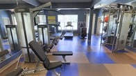 This fitness center offers a focused and functional workout environment designed to support a range of exercise routines. The space is finished with striking navy blue walls that create an energetic atmosphere, complemented by a distinctive two-tone rubber flooring in blue and tan — a practical and visually dynamic choice that defines separate workout zones.

Cardio equipment, including two treadmills and an elliptical machine, is positioned near the windows along the far wall, allowing users to exercise while benefiting from natural light and exterior views of surrounding greenery. A multi-station cable machine and an adjustable weight bench are prominently featured toward the front of the space, catering to strength training needs.

Large mirrors line one wall, enhancing the sense of openness and allowing users to monitor their form during workouts. Exercise balls and additional accessories are visible along the opposite side, adding versatility to available training options. Overhead lighting ensures the space remains bright and well-illuminated throughout. The overall layout is thoughtfully organized, maximizing usability while maintaining an open, uncluttered feel.