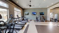 Located at 5501 Old Montgomery Hwy in Tuscaloosa, Alabama, this fitness center is a thoughtfully designed, single-floor facility offering a comprehensive workout environment. The space is organized into distinct functional zones, seamlessly connected by a striped, multi-tone flooring pattern that guides movement throughout the room.

The cardio area is lined with a row of TRUE treadmills and elliptical trainers, all positioned to take advantage of the large, multi-pane windows that flood the space with natural light and provide views of the surrounding exterior. Ceiling fans are strategically placed throughout to maintain comfortable airflow during workouts.

The strength training section features a variety of cable and selectorized machines, along with a well-stocked free weight area that includes an organized dumbbell rack, kettlebell rack, and a barbell/curl bar rack — catering to a wide range of training styles. Multiple flat-screen TVs are mounted along the walls, keeping members engaged throughout their sessions.

The facility is accessed through a set of elegant black-framed glass double doors that open to an adjacent lounge or common area, adding a polished, upscale aesthetic to the overall design. Crown molding, recessed lighting, and a neutral sage-toned color palette contribute to a clean, modern ambiance that balances functionality with refined style.