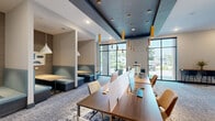 Located at 554 Lancaster Ave in Malvern, Pennsylvania, this contemporary coworking space offers a thoughtfully designed, single-floor layout that balances collaborative and private work environments. The open-plan main area features long communal workstations equipped with desktop computers and privacy dividers, illuminated by striking gold pendant lights that hang from a bold teal coffered ceiling — a defining architectural element that anchors the space. Patterned carpeting in neutral tones adds texture throughout, complementing the modern, professional aesthetic. Floor-to-ceiling windows along the perimeter flood the interior with abundant natural light and provide open views of the surrounding greenery, enhancing the sense of spaciousness. A lounge area with teal upholstered booth seating offers a comfortable alternative for casual work or informal meetings. The space also includes at least two enclosed conference rooms, each featuring a wall-mounted flat-screen display, textured accent walls with a mosaic-style tile pattern, a central wood conference table, and leather rolling chairs. One conference room benefits from direct access to natural light through adjacent glass walls. A glass-panel partition elegantly separates the private conference room from the open workspace, maintaining visual connectivity while ensuring privacy. The overall ambiance is polished, functional, and design-forward.