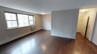 This single-floor space offers a clean, straightforward layout designed for functional living. The main living area immediately draws attention with its rich, dark-stained hardwood floors that run throughout, adding warmth and character to the otherwise neutral palette. Walls are finished in a soft gray tone, complemented by crisp white baseboards and trim that lend a clean, modern contrast.

Natural light enters through a double-hung window, which also provides views of the surrounding urban streetscape. Beneath the window sits a wall-mounted air conditioning unit paired with a baseboard heating system running along the perimeter, ensuring year-round climate comfort. A flush-mount ceiling light fixture provides overhead illumination and maintains the room's understated aesthetic.

The open floor plan allows for flexible furniture arrangement, making the space adaptable to a variety of needs. An accent wall in a slightly deeper gray shade adds subtle depth and visual interest to the room, helping to define different zones within the open layout. The overall ambiance is calm and neutral, providing a blank canvas well-suited for personalization. The space strikes a balance between urban practicality and residential comfort, making it a functional and inviting environment.