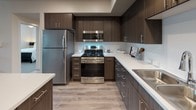 This thoughtfully designed 2-bedroom, 2-bathroom residence offers a contemporary living experience across a single level. The open-concept kitchen is a standout feature, showcasing rich dark wood cabinetry with sleek chrome hardware, bright white quartz countertops, a clean tile backsplash, and a full suite of stainless steel appliances including a gas range, built-in microwave, refrigerator, and dishwasher. Light wood-style flooring runs throughout the main living areas, complementing the warm, neutral color palette. The living area provides a comfortable and stylish space with room for relaxed seating and decor. Both bedrooms are carpeted and filled with natural light from large windows with views of lush greenery. The primary bathroom features a long double-sink vanity with dark wood cabinetry and a generous mirror, while the second bathroom offers a similarly styled vanity paired with a shower-tub combo. A dedicated in-unit laundry area houses side-by-side washer and dryer units with a built-in shelf above. The residence also includes an attached private garage with an automatic opener, offering convenient and secure parking. The overall design balances modern aesthetics with functional living spaces.
