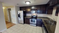 This single-floor space includes a well-appointed kitchen that serves as a functional centerpiece. The kitchen features rich, dark espresso-toned cabinetry extending from floor to ceiling, providing generous storage capacity through both upper and lower cabinets. The cabinetry is complemented by granite countertops with subtle speckled patterning, adding a polished, durable work surface throughout.

Stainless steel appliances outfit the kitchen, including a side-by-side refrigerator with an in-door ice and water dispenser, an electric range with oven, an over-the-range microwave, and what appears to be a dishwasher. The appliances create a cohesive, modern aesthetic against the darker cabinetry.

The flooring consists of light-toned ceramic tile laid in a classic grid pattern, brightening the space and offering easy maintenance. Recessed ventilation and a flush-mount ceiling light fixture provide functional overhead illumination. The neutral wall tones contrast cleanly with the dark cabinetry, contributing to a balanced, contemporary interior feel throughout the kitchen area.