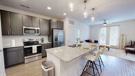 Located in Granbury, TX, this 2-bedroom, 2-bathroom residence offers a thoughtfully designed single-floor layout. The open-concept kitchen and living area serves as the heart of the home, where wood-style flooring flows beneath a kitchen island topped with granite countertops and illuminated by elegant pendant lighting. Dark cabinetry, a decorative tile backsplash, and a full suite of stainless steel appliances — including a refrigerator, range, and microwave — complete the kitchen's modern aesthetic. The adjoining living room features plush carpeting, a ceiling fan, and French doors that open to a private balcony, inviting abundant natural light and outdoor views. The primary bedroom is a comfortable retreat with a bright window, ceiling fan, and direct access to its en-suite bathroom, which boasts a wide granite vanity with dual sinks, dark wood cabinetry, and a glass-enclosed shower. The second bedroom is cozy and well-lit, complemented by a second full bathroom with matching finishes. Both bedrooms are served by dedicated walk-in closets featuring built-in shelving and hanging rods, offering generous storage throughout. Clean white walls and consistent, contemporary finishes create a cohesive and inviting atmosphere throughout the entire space.