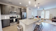 Located in Granbury, TX, this 2-bedroom, 2-bathroom residence offers a thoughtfully designed single-floor layout. The open-concept kitchen and living area serves as the heart of the home, where wood-style flooring flows beneath a kitchen island topped with granite countertops and illuminated by elegant pendant lighting. Dark cabinetry, a decorative tile backsplash, and a full suite of stainless steel appliances — including a refrigerator, range, and microwave — complete the kitchen's modern aesthetic. The adjoining living room features plush carpeting, a ceiling fan, and French doors that open to a private balcony, inviting abundant natural light and outdoor views. The primary bedroom is a comfortable retreat with a bright window, ceiling fan, and direct access to its en-suite bathroom, which boasts a wide granite vanity with dual sinks, dark wood cabinetry, and a glass-enclosed shower. The second bedroom is cozy and well-lit, complemented by a second full bathroom with matching finishes. Both bedrooms are served by dedicated walk-in closets featuring built-in shelving and hanging rods, offering generous storage throughout. Clean white walls and consistent, contemporary finishes create a cohesive and inviting atmosphere throughout the entire space.