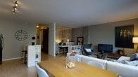 This single-floor residence offers a thoughtfully designed open-concept layout that seamlessly connects the dining and living areas into one cohesive, functional space. The dining area is anchored by a warm wooden table surrounded by light upholstered chairs, with a simple glass centerpiece adding a refined touch. Adjacent to the dining space, the living area is furnished with a comfortable cream sofa and a sculptural accent chair in charcoal, contributing to a sophisticated, contemporary palette. A standout feature of the space is the decorative mirrored panel wall, which reflects ambient light and adds depth and elegance to the interior. A dedicated workspace or console area is tucked behind a low dividing unit, illuminated by warm task lighting that creates an inviting nook. Shelving units provide practical storage while maintaining the open feel of the layout. The walls are finished in a cool grey tone, complemented by smooth ceilings and light hardwood flooring that runs throughout, lending warmth and continuity to the space. A concrete structural column adds an industrial character that contrasts beautifully with the polished furnishings. Overall, the space strikes a balance between modern design and everyday comfort.