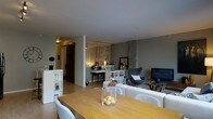 This single-floor residence offers a thoughtfully designed open-concept layout that seamlessly connects the dining and living areas into one cohesive, functional space. The dining area is anchored by a warm wooden table surrounded by light upholstered chairs, with a simple glass centerpiece adding a refined touch. Adjacent to the dining space, the living area is furnished with a comfortable cream sofa and a sculptural accent chair in charcoal, contributing to a sophisticated, contemporary palette. A standout feature of the space is the decorative mirrored panel wall, which reflects ambient light and adds depth and elegance to the interior. A dedicated workspace or console area is tucked behind a low dividing unit, illuminated by warm task lighting that creates an inviting nook. Shelving units provide practical storage while maintaining the open feel of the layout. The walls are finished in a cool grey tone, complemented by smooth ceilings and light hardwood flooring that runs throughout, lending warmth and continuity to the space. A concrete structural column adds an industrial character that contrasts beautifully with the polished furnishings. Overall, the space strikes a balance between modern design and everyday comfort.