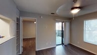 This single-floor residence offers a clean and functional layout designed for comfortable everyday living. The main living area features rich, dark hardwood-style flooring that extends throughout, providing a warm and cohesive aesthetic. Neutral-toned walls complement the space, creating a bright and versatile canvas. A ceiling fan is centrally positioned, adding both practicality and a subtle design element to the room.

One of the standout features of the living area is the full-height sliding glass door, dressed with vertical blinds, which opens to a private outdoor balcony or patio. This connection to the outdoors allows for natural light to fill the space while offering a seamless transition to the exterior.

An open doorway leads to an adjoining room, also finished with matching dark hardwood-style floors and its own ceiling fan, suggesting a well-connected and consistent interior design throughout. The textured ceiling adds a layer of dimension to each room. Overall, the space presents a practical and inviting atmosphere, with thoughtful details such as updated flooring, ample wall outlets, and a layout that balances openness with defined living zones.