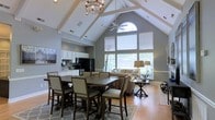 This single-floor space makes a striking first impression with its dramatic vaulted ceilings adorned with exposed white beams that draw the eye upward and create an expansive, airy atmosphere. An elegant chandelier and a ceiling fan complement the architectural grandeur, while a large arched window floods the space with abundant natural light, enhancing the open and welcoming ambiance.

The open-concept layout seamlessly integrates the dining area, living room, and kitchen into one cohesive living space. The dining area is anchored by a substantial dark wood farmhouse-style table surrounded by upholstered chairs, set atop a patterned area rug that adds texture and warmth to the hardwood floors. Adjacent to the dining space, the living area is thoughtfully furnished with comfortable seating arranged to take full advantage of the natural light and outdoor views provided by the large windows.

The kitchen features white cabinetry, black appliances, and ample counter space, offering both functionality and a clean aesthetic that complements the overall neutral color palette of soft grays and warm wood tones. Recessed lighting throughout further enhances the brightness of the space. The overall design balances casual comfort with refined style, making this a versatile and inviting environment suited for both everyday living and entertaining.