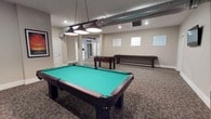 This spacious game room is designed for recreation and social engagement, offering a well-curated selection of classic games in a comfortable, open setting. The centerpiece of the room is a full-size pool table with a vibrant teal felt surface, illuminated by a trio of pendant lights suspended from chains — a stylish focal point that draws the eye immediately upon entering. Beyond the pool table, a foosball table and a regulation-length shuffleboard table provide additional entertainment options, making the space suitable for a wide range of guests and activities. The room features neutral beige walls and patterned carpeted flooring that adds warmth and helps define the recreational atmosphere. Exposed silver ductwork runs across the ceiling, lending an industrial character that contrasts attractively with the otherwise clean, modern finishes. Recessed lighting complements the pendant fixtures, ensuring the space is well-lit throughout. Small horizontal windows along one wall allow modest natural light to filter in, while a glass-paned door at the rear connects the room to adjacent areas, contributing to an open and accessible feel.