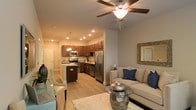 This single-floor residence showcases an open-concept design that seamlessly connects the living, dining, and kitchen areas into one cohesive and functional space. The living area is centered around a comfortable neutral-toned sofa accented with blue throw pillows, complemented by a glass-top coffee table and a decorative framed mirror on the wall. A ceiling fan with a built-in light fixture provides both airflow and ambient lighting overhead.

The kitchen is a standout feature, outfitted with rich, warm-toned cabinetry, granite countertops, and a full suite of stainless steel appliances, including a refrigerator, stove, and microwave. Pendant lighting above the kitchen peninsula adds a refined touch while enhancing task lighting. Recessed ceiling lights throughout ensure the space is well-illuminated.

Light wood-tone flooring runs continuously across the open floor plan, contributing to the warm and unified aesthetic. A dining area is positioned near the entrance, offering a natural transition between spaces. The overall ambiance is modern yet approachable, balancing practical functionality with stylish, contemporary finishes.