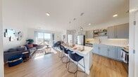 This well-appointed single-floor residence showcases a thoughtfully designed open-concept layout that seamlessly connects the kitchen, dining, and living areas. The kitchen is a standout feature, outfitted with soft gray shaker cabinets, white subway tile backsplash, stainless steel appliances — including a refrigerator, range, microwave, and dishwasher — and open shelving for added storage and display. A large central island with a white quartz-style countertop and seating for multiple bar-height chairs serves as both a functional workspace and a casual dining hub, illuminated by a trio of modern pendant lights overhead. Light wood-style flooring flows throughout the main living areas, enhancing the bright and airy atmosphere. The living room is generously sized and bathed in natural light from floor-to-ceiling sliding glass doors that open to a private balcony, offering elevated outdoor views of the surrounding cityscape. The two bedrooms are carpeted for comfort and each features a distinctive accent wall — one with a textured script-style wallcovering and the other with a soft neutral textile-look finish. The bathroom includes a full soaking tub with a white tile surround and brushed gold fixtures. Recessed lighting and clean white walls throughout contribute to the space's modern, cohesive aesthetic.