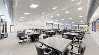 Located at 2215 Hailey Dr in Marlton, NJ, this single-floor community recreation center offers a generously sized, open-plan layout designed to accommodate a wide range of social and leisure activities. The space is defined by its clean, modern aesthetic, featuring a neutral color palette of whites and grays throughout, with light-toned carpet flooring and a drop-tile ceiling outfitted with recessed panel lighting that ensures bright, even illumination across the entire floor.

The main hall is expansive, accommodating numerous game and activity tables with rolling chairs arranged throughout, suitable for card games, board games, and group gatherings. A prominent wall inscription reading "Gather, Play, Enjoy!" reinforces the space's welcoming and communal purpose. Large windows positioned along the upper walls allow natural light to filter in while offering views of the surrounding greenery, enhancing the open and airy atmosphere. An oversized ceiling fan adds to the comfort of the space.

Recreational amenities include a full-size ping-pong table, a shuffleboard table, and a pool table, catering to a variety of interests. A lounge area with comfortable seating and a wall-mounted flat-screen television provides a relaxed setting for casual gatherings. A secondary area features additional lounge seating, colorful artwork, and open floor space for flexible use. Exposed ductwork and industrial-style elements in select areas add architectural character to this vibrant, activity-focused community space.