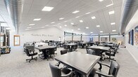 Located at 2215 Hailey Dr in Marlton, NJ, this single-floor community recreation center offers a generously sized, open-plan layout designed to accommodate a wide range of social and leisure activities. The space is defined by its clean, modern aesthetic, featuring a neutral color palette of whites and grays throughout, with light-toned carpet flooring and a drop-tile ceiling outfitted with recessed panel lighting that ensures bright, even illumination across the entire floor.

The main hall is expansive, accommodating numerous game and activity tables with rolling chairs arranged throughout, suitable for card games, board games, and group gatherings. A prominent wall inscription reading "Gather, Play, Enjoy!" reinforces the space's welcoming and communal purpose. Large windows positioned along the upper walls allow natural light to filter in while offering views of the surrounding greenery, enhancing the open and airy atmosphere. An oversized ceiling fan adds to the comfort of the space.

Recreational amenities include a full-size ping-pong table, a shuffleboard table, and a pool table, catering to a variety of interests. A lounge area with comfortable seating and a wall-mounted flat-screen television provides a relaxed setting for casual gatherings. A secondary area features additional lounge seating, colorful artwork, and open floor space for flexible use. Exposed ductwork and industrial-style elements in select areas add architectural character to this vibrant, activity-focused community space.