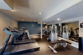 Located at 11402 Evans St in Omaha, Nebraska, this well-appointed fitness center offers a comprehensive workout facility across a single floor. The main exercise area is generously sized, featuring durable rubber flooring, a textured ceiling with recessed lighting, and structural columns that define the space without interrupting the open layout. The workout floor is equipped with multiple treadmills, a stair climber, a full dumbbell rack, and a selection of strength training machines, with a large wall mirror enhancing the sense of space and allowing users to monitor their form. A bold blue accent wall provides a modern, energizing aesthetic contrast to the neutral beige walls throughout. Natural light filters in through several windows, offering views of the surrounding outdoor area. Adjacent to the main workout floor is a tiled restroom corridor featuring clearly marked, ADA-accessible men's and women's facilities. The entry area includes a carpeted reception zone with a first aid kit, an AED station, a vending machine, and a pass-through window, ensuring convenience and safety for all users. The overall ambiance is functional and motivating, designed to support a well-rounded fitness experience.