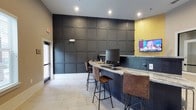 This well-appointed single-floor amenity space blends modern design with inviting functionality. The reception and lounge area welcomes visitors with warm neutral tones, exposed brick accent walls, pendant lighting, and comfortable seating accented with colorful throw pillows. Framed butterfly artwork adds a decorative touch, while a business center workstation tucked nearby provides a practical workspace complete with a desktop computer and printer.

A curved granite-topped bar counter serves as a central hub, surrounded by leather-upholstered bar stools with sleek metal frames. The counter area is equipped with computer workstations and a wall-mounted flat-screen TV, set against a dramatic floor-to-ceiling dark paneled accent wall with a bold geometric grid pattern. A gold-toned accent wall adds a warm contrast to the otherwise cool, sophisticated palette.

The game and recreation area is highlighted by a full-size billiards table bathed in natural light from large bay windows, creating an open and airy atmosphere. Adjacent to this, a dedicated game zone features a shuffleboard table against a dark brick-effect accent wall boldly emblazoned with the phrase "LET'S PLAY!" Throughout the space, large-format light stone tile flooring and herringbone-patterned inlays tie the various zones together cohesively.